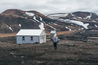 Ein Wanderer läuft auf eine kleine Berghütte in einer dunklen Vulkanlandschaft mit Schneefeldern zu