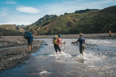 Wanderer mit Trekkingstöcken waten durch einen flachen Gletscherfluss in einem steinigen Tal